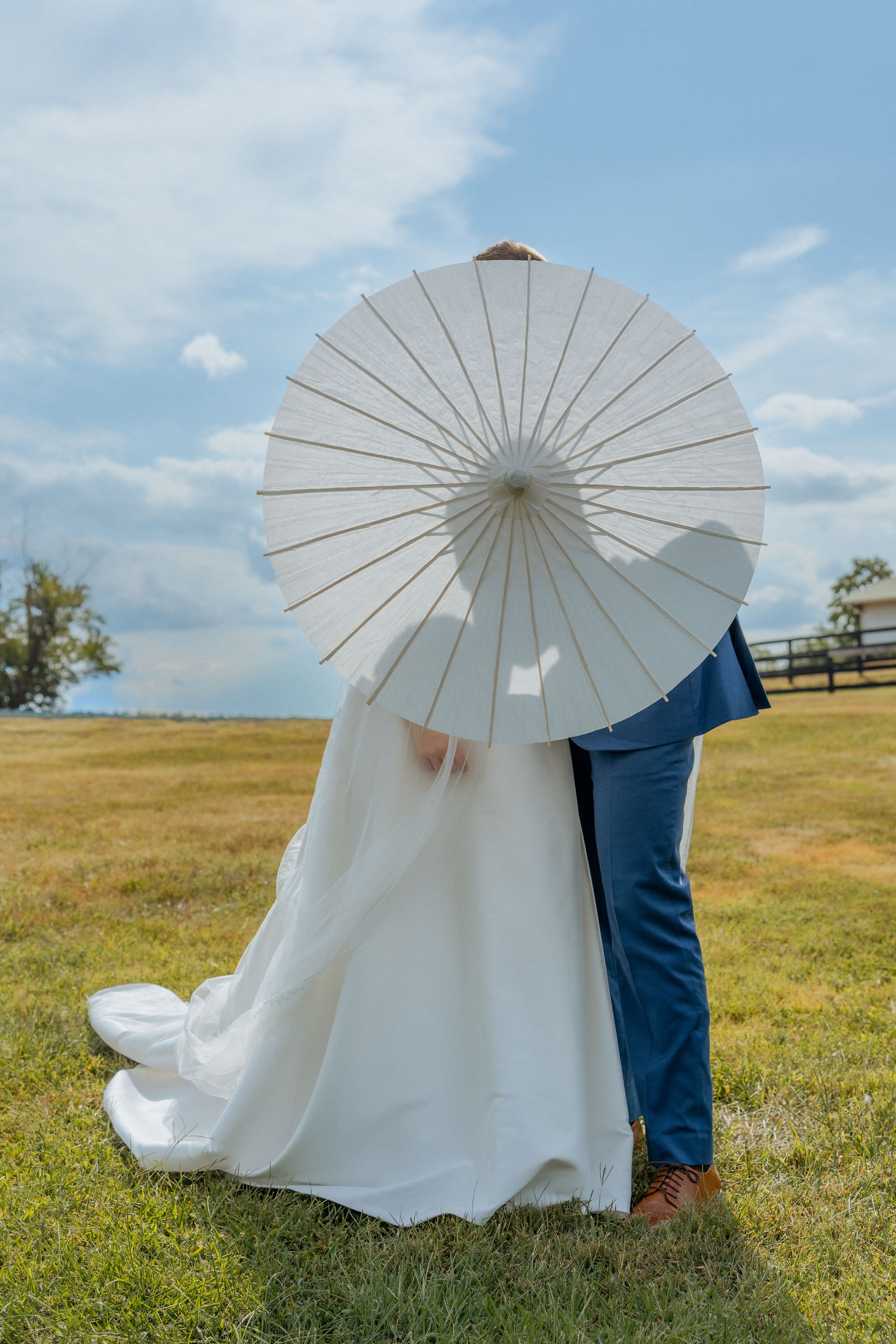 A bride and groom pose together under a white parasol in an open grassy field on a sunny day.