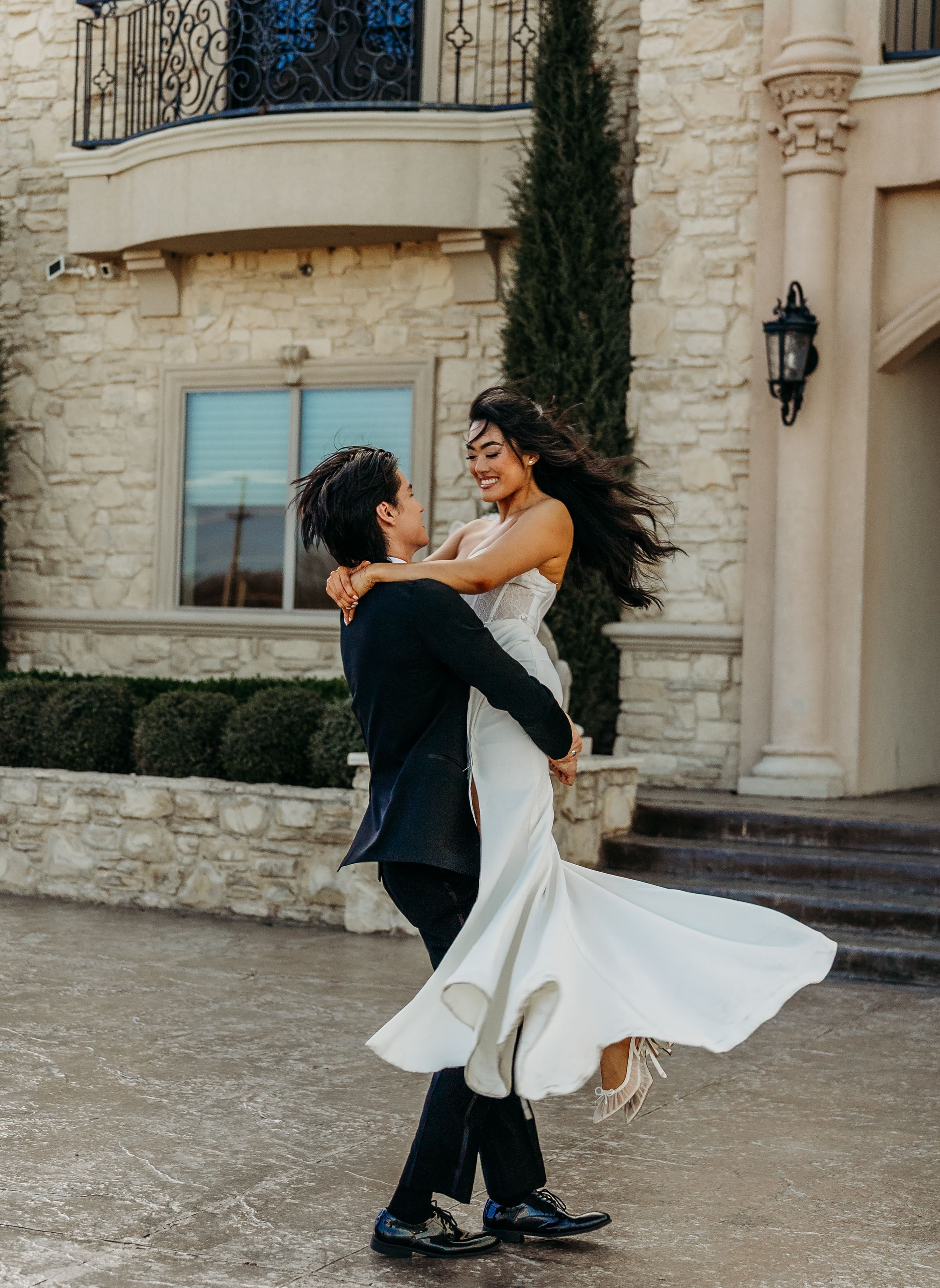 A groom in a black suit lifts and twirls his bride in a white flowing dress in front of an elegant stone building with Mediterranean architecture.