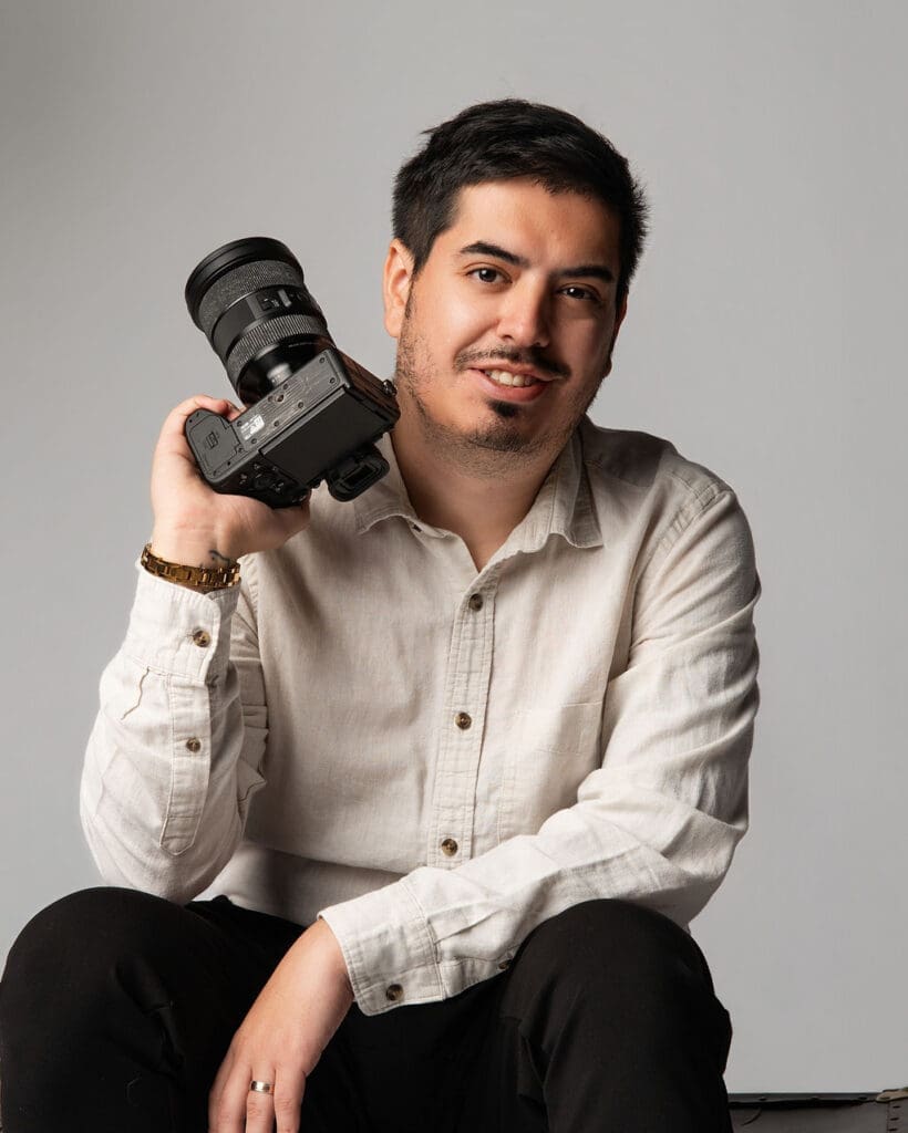 A professional photographer poses for a portrait while holding a DSLR camera, wearing a light-colored button-up shirt against a neutral gray studio background.