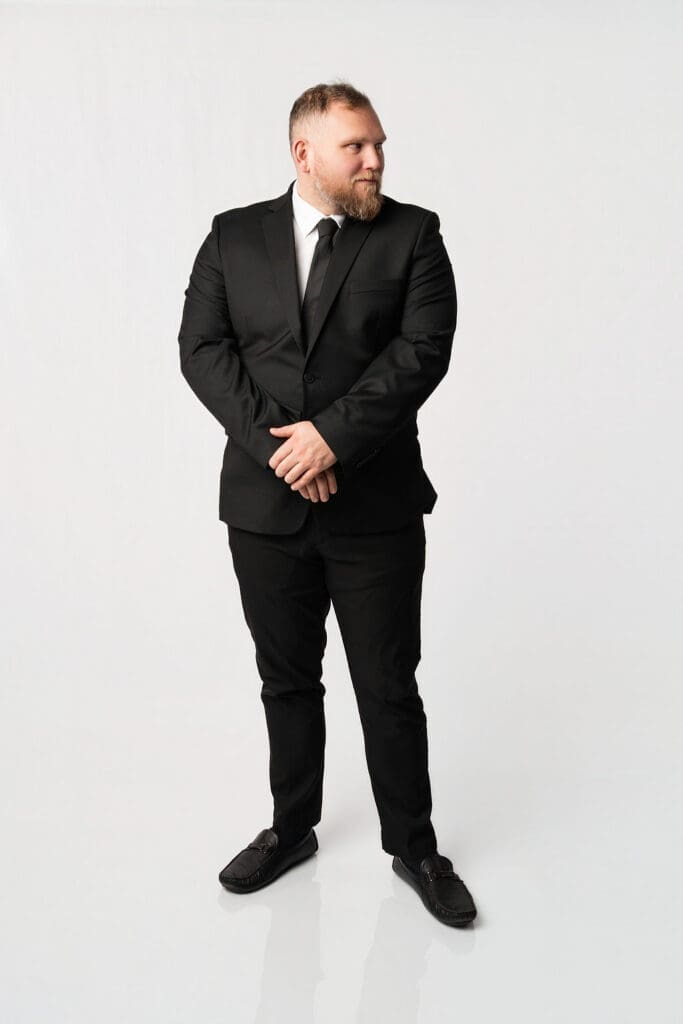 A professional man in a formal black suit stands against a plain white background in a studio portrait setting.