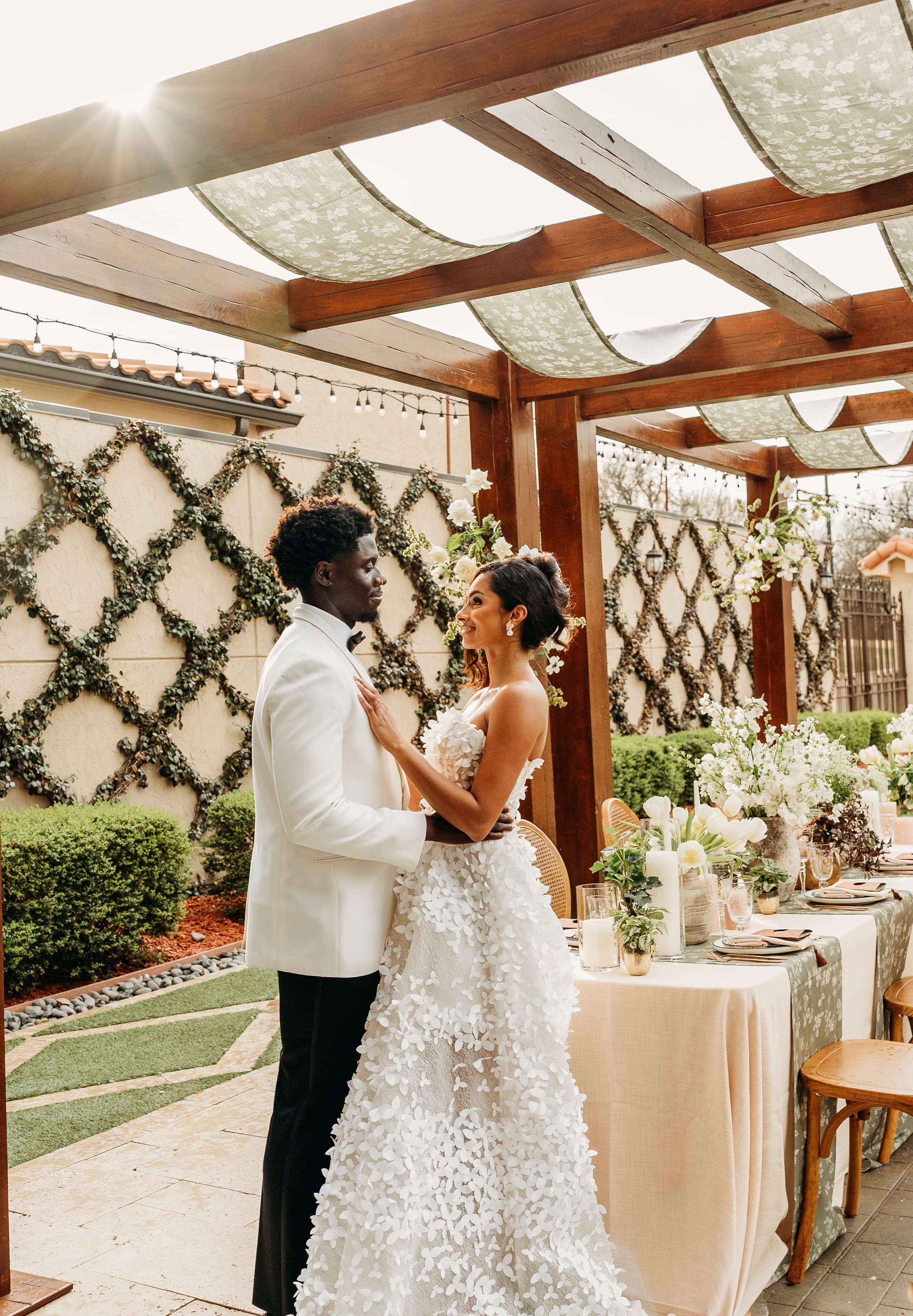 A bride and groom embrace under a wooden pergola with decorative geometric backdrop during their wedding ceremony in a bright, sunlit venue.