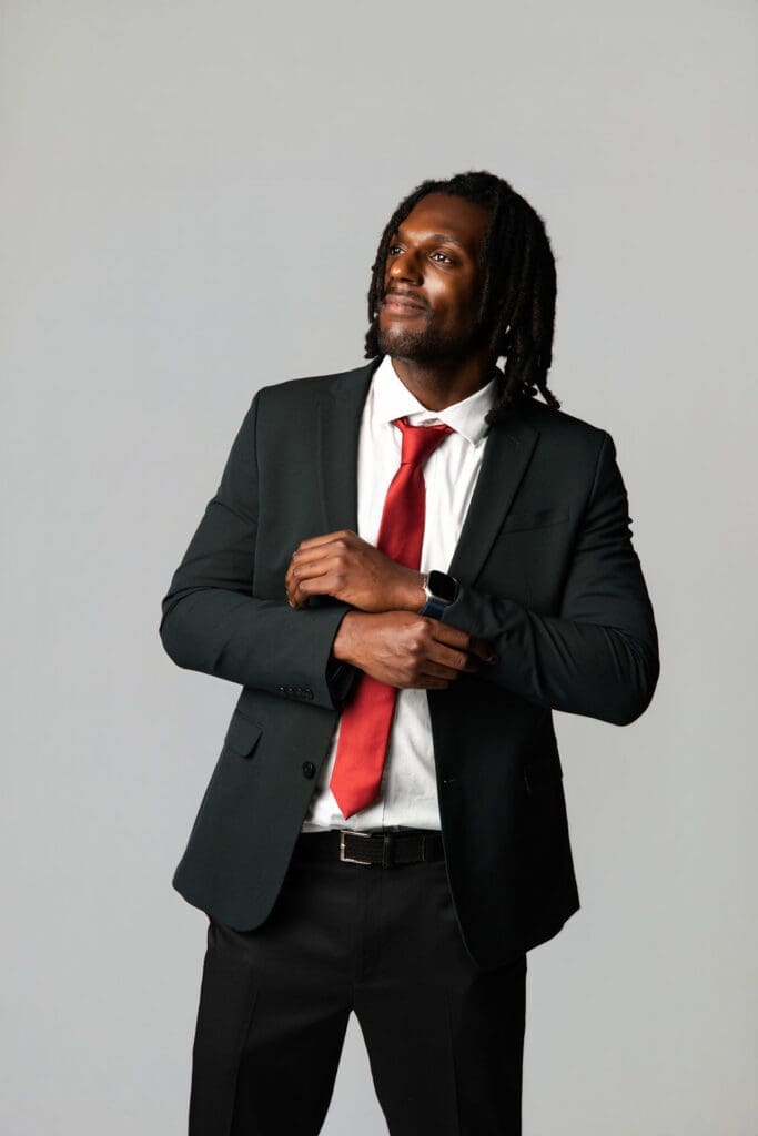 A professional man in a dark business suit and red tie adjusts his jacket against a neutral white background.