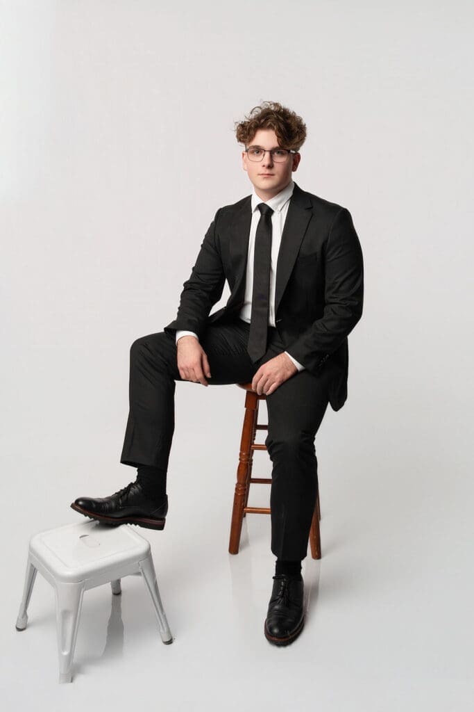 A young man in formal business attire sits on a wooden stool against a plain white background in a professional studio portrait.