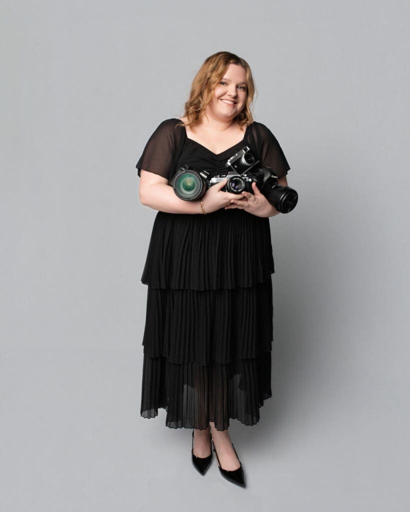 A woman wearing a black tiered midi dress and black shoes stands against a neutral gray background in a studio portrait setting.