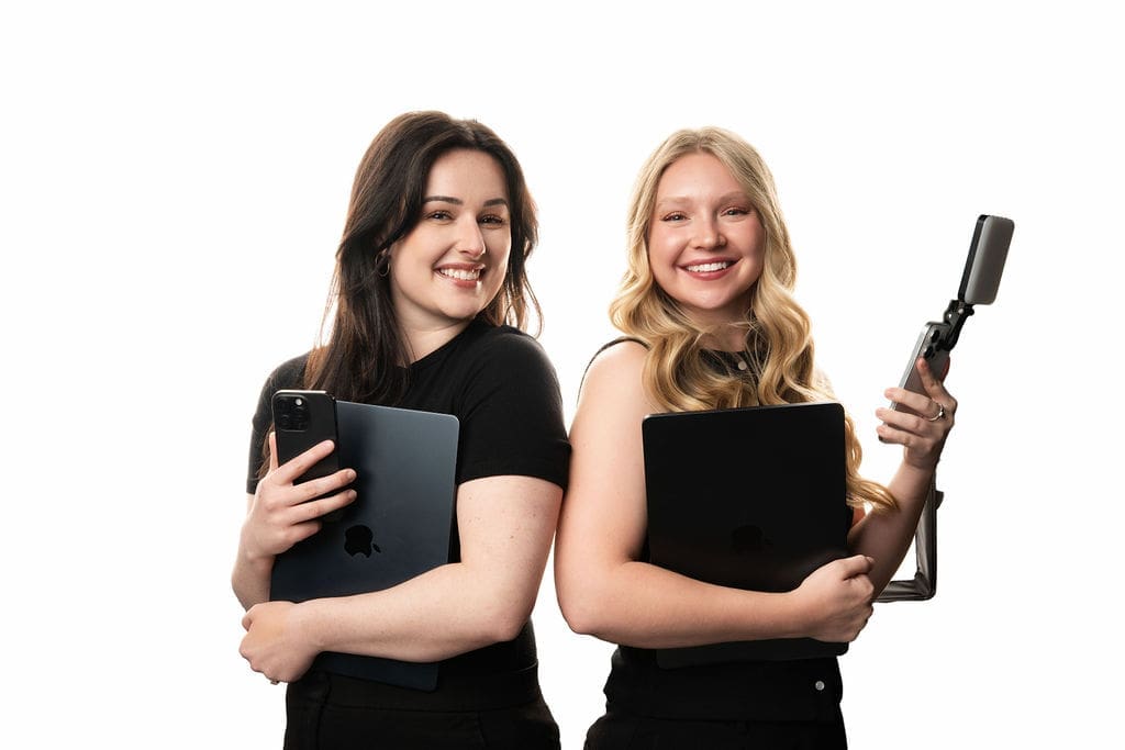 Two young women in black graduation attire pose together holding diplomas and academic caps against a white background.