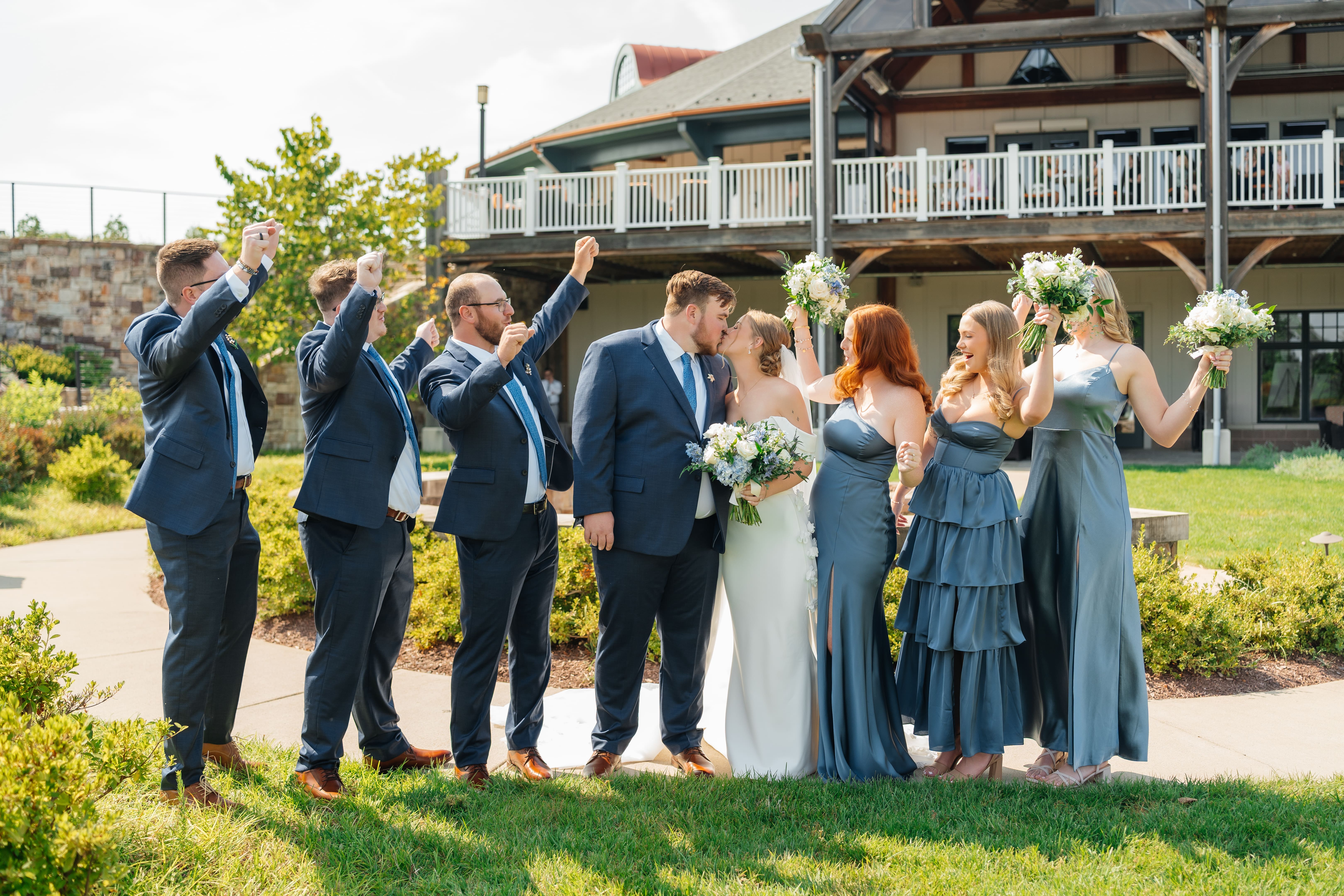 A wedding party of approximately 10 people stands in a garden in front of a large house, with the bride in white and attendants in navy blue dresses and suits.