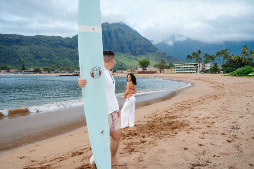 A woman in a white dress stands on a sandy beach holding a white surfboard with mountains and palm trees visible in the background.