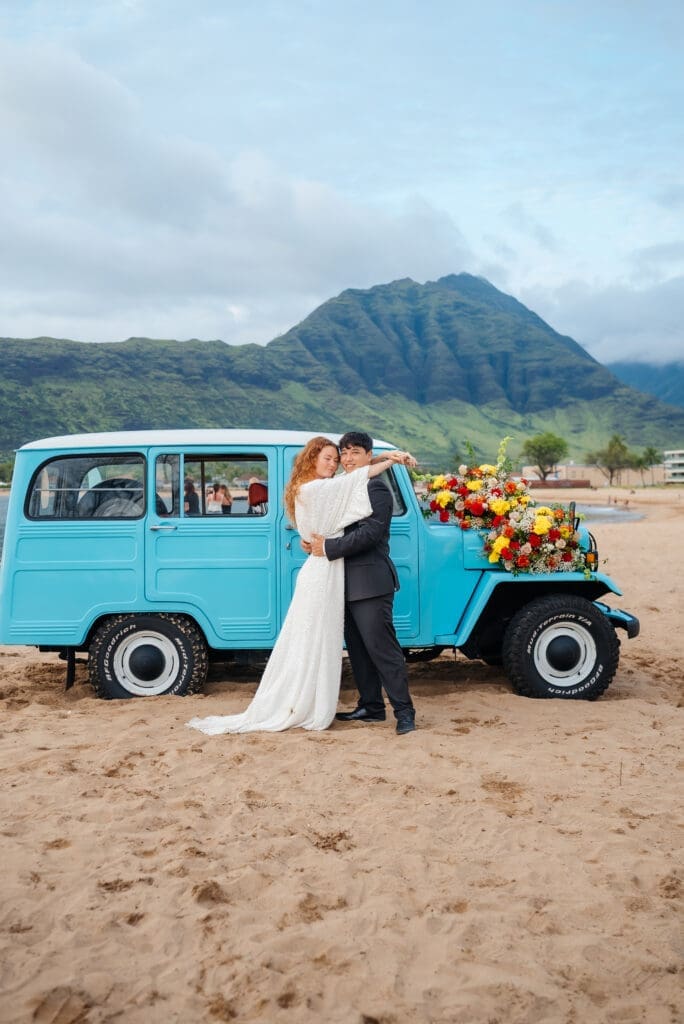 A bride and groom pose together beside a turquoise vintage van decorated with flowers on a sandy beach with mountains in the background.
