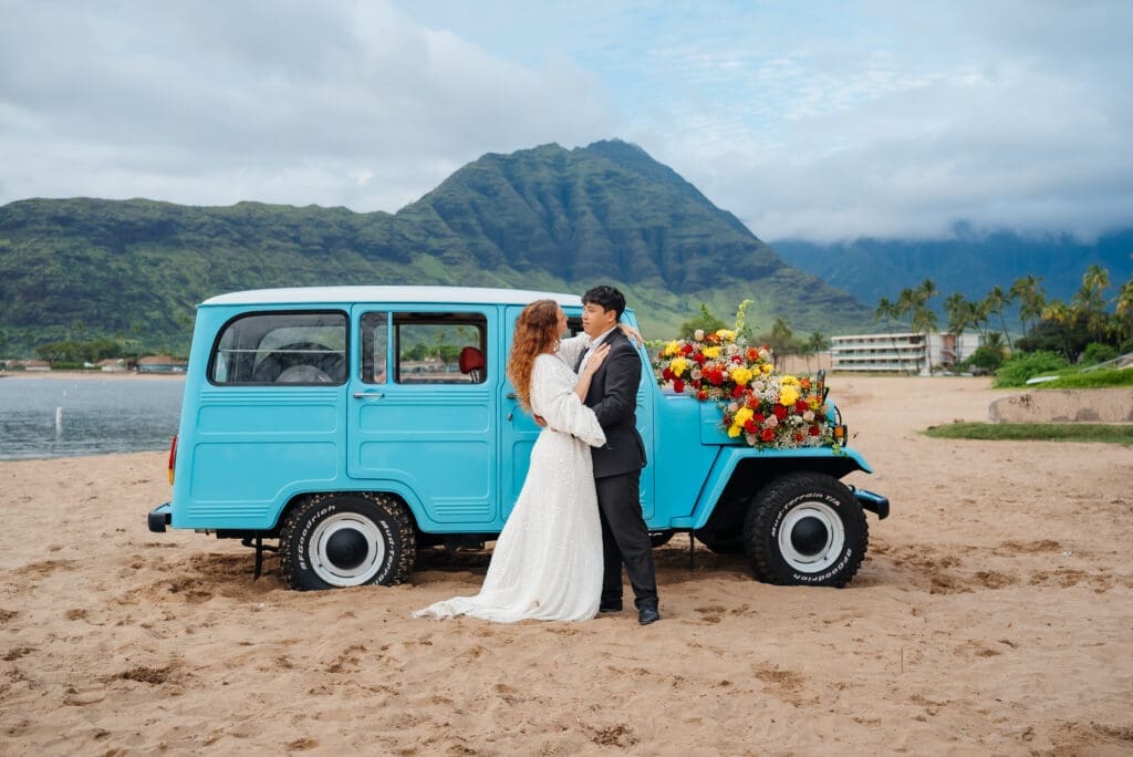 A bride and groom pose together beside a vintage turquoise truck decorated with flowers on a sandy beach with mountains in the background.