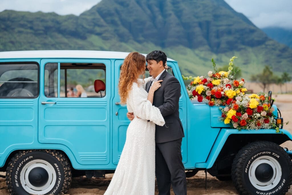 A bride and groom kiss beside a turquoise vintage truck decorated with colorful flowers, with a dramatic mountain landscape in the background.