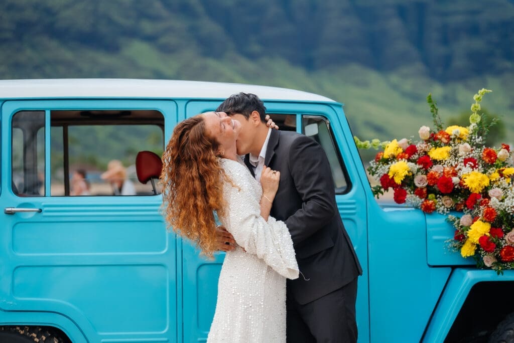 A bride and groom kiss beside a turquoise vintage truck decorated with colorful flowers in an outdoor wedding celebration.