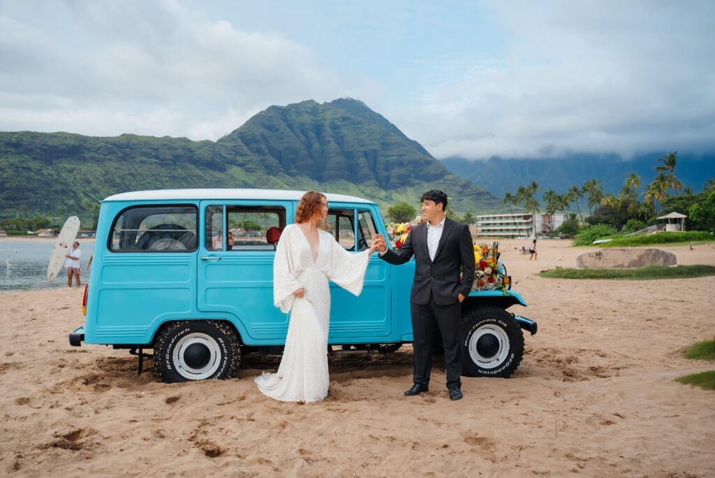 A bride and groom pose together beside a charming turquoise vintage truck on a sandy beach with mountains in the background.