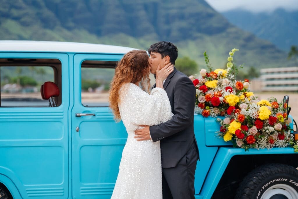 A bride and groom share a kiss next to a vintage turquoise truck decorated with colorful flowers in a scenic mountain countryside setting.