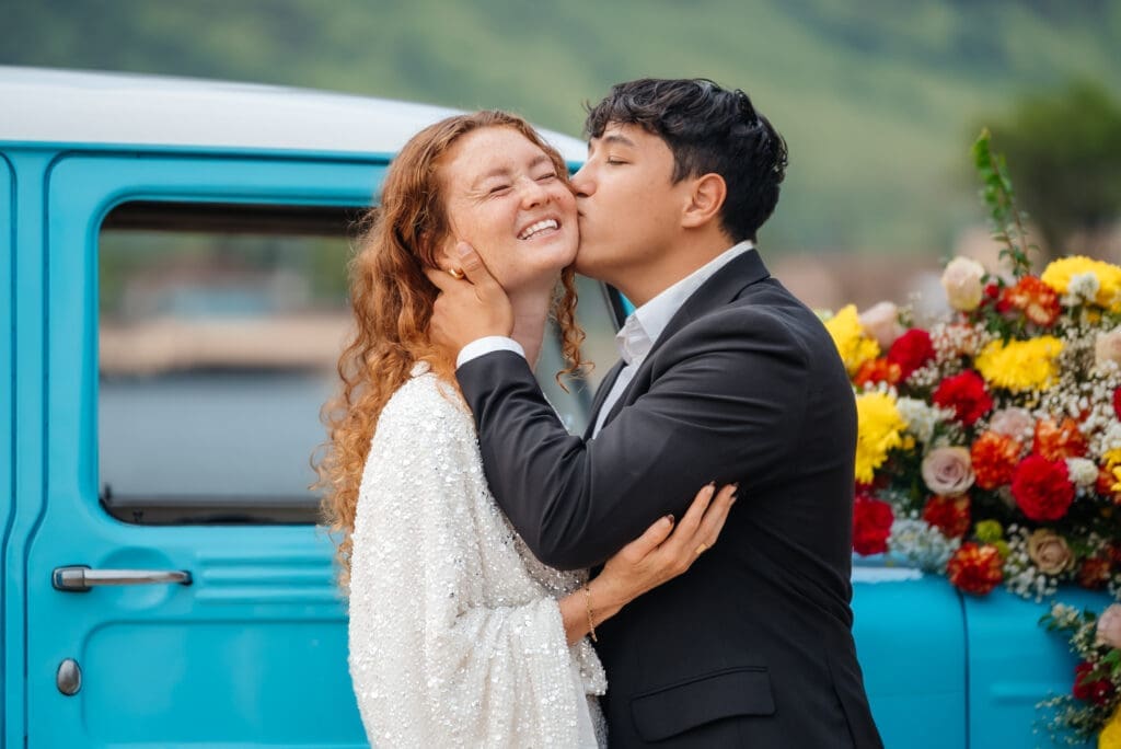 A newlywed couple shares a kiss in front of a vintage turquoise truck decorated with colorful flowers, capturing a romantic wedding moment outdoors.