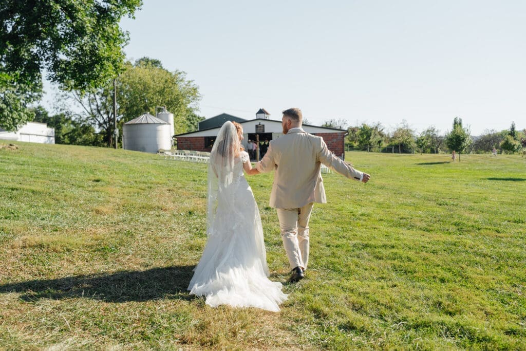 A bride and groom walk hand-in-hand through a sprawling green field on their wedding day, with a farmhouse visible in the background.
