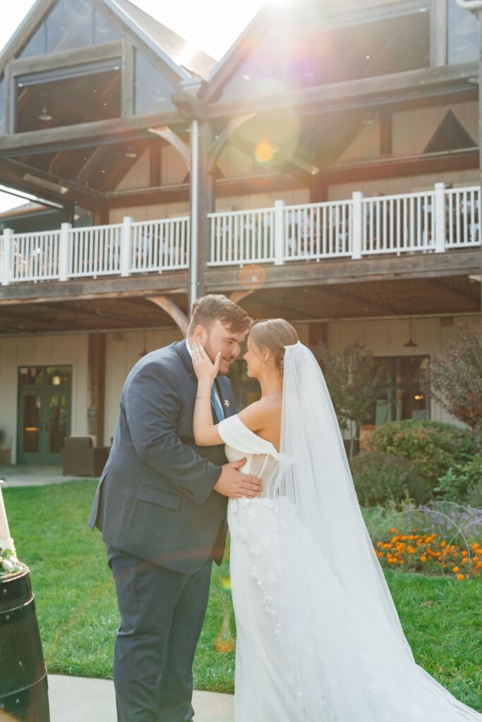 A bride and groom share a kiss on the lawn in front of an elegant mansion during golden hour, with blooming flowers and manicured grounds visible in the background.