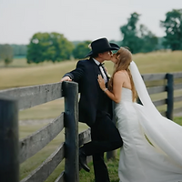 A bride and groom share an intimate kiss by a wooden fence in a rural field during golden hour.