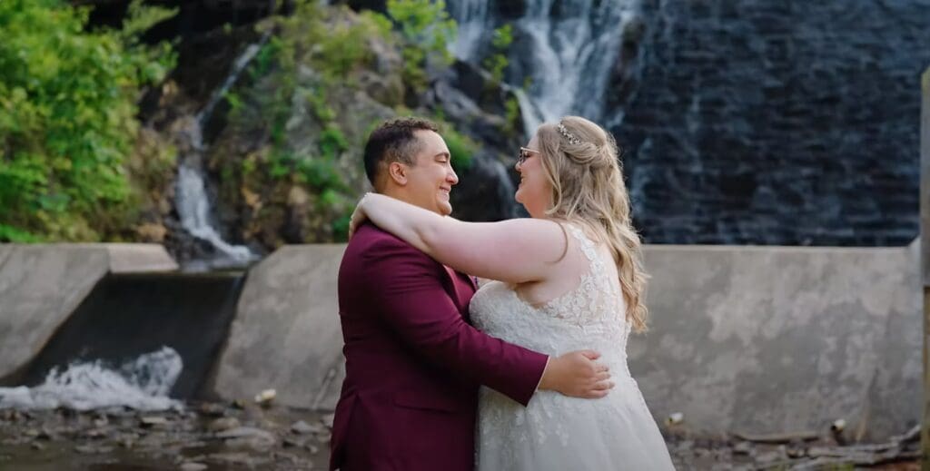 A bride and groom embrace and kiss during their wedding photo session by a stone wall and waterfall backdrop.