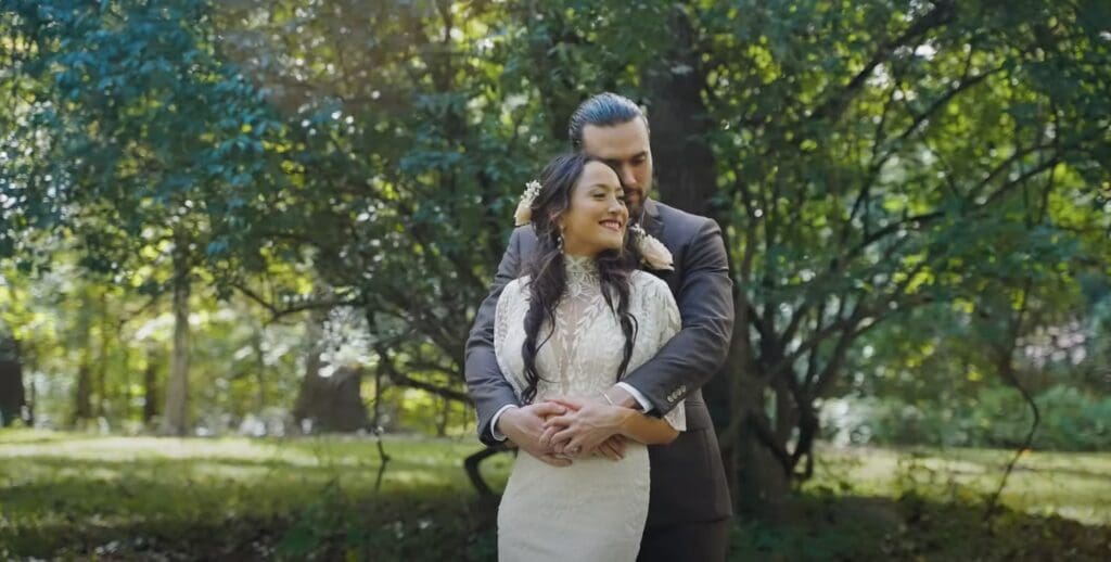 A newlywed couple poses together in a lush park setting during golden hour, with the bride in a white dress and groom in a dark suit surrounded by green foliage.