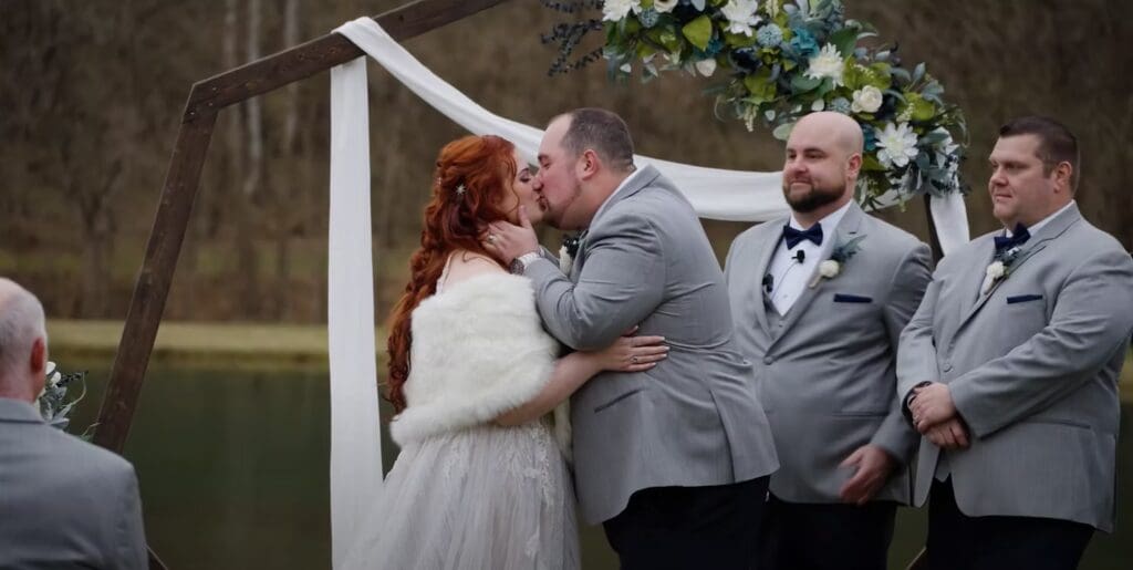 A bride and groom share a kiss during their wedding ceremony at an outdoor venue with groomsmen standing nearby and a decorated white arch in the background.