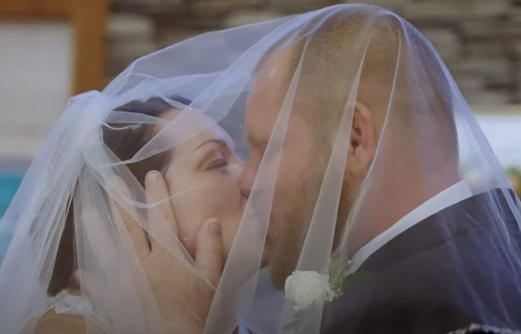 A bride and groom share a romantic kiss beneath delicate white tulle netting during their wedding ceremony.
