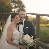 A bride and groom pose together outdoors during golden hour, with the bride holding a white bouquet and wearing a lace wedding dress and veil.