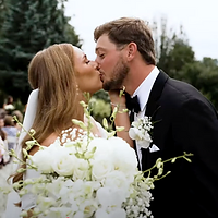 A bride and groom share a kiss at their wedding ceremony surrounded by white floral arrangements with guests visible in the background.
