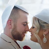 A bride and groom share a romantic kiss at their wedding ceremony, both wearing formal attire with the bride in a white hat and holding a red rose.