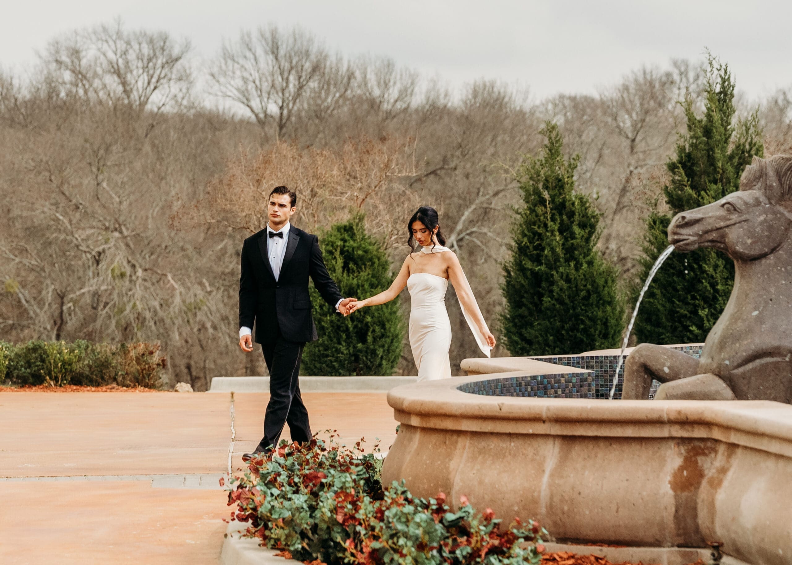 Bride and groom holding hands at a stone fountain in a formal garden
