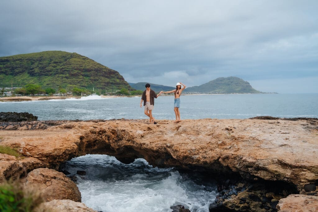 Two people wade in shallow water on a rocky beach with volcanic islands visible in the distance under a partly cloudy sky.