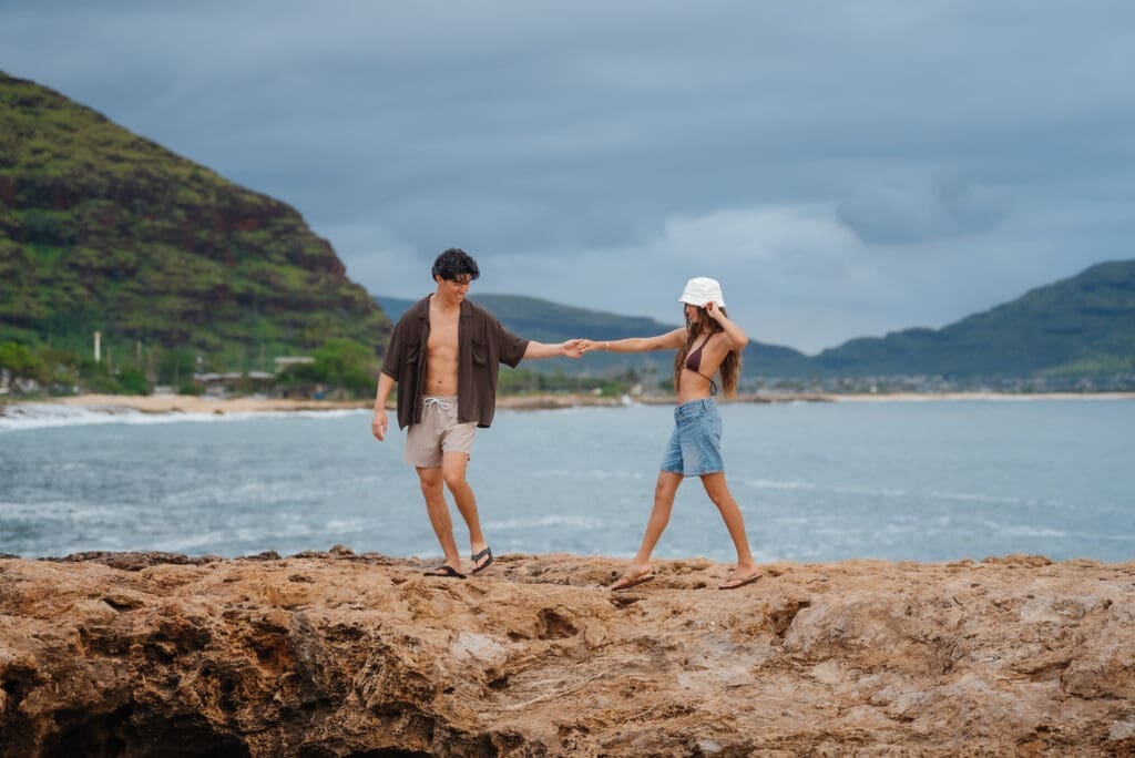 Two people engage in beach activities on a sandy shore with mountains and ocean visible in the background under a partly cloudy sky.