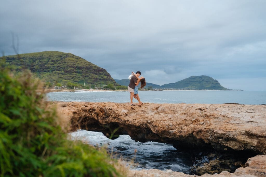 Two hikers walk along a pristine sandy beach with weathered driftwood in the foreground, surrounded by dramatic coastal cliffs and distant islands under a partly cloudy sky.
