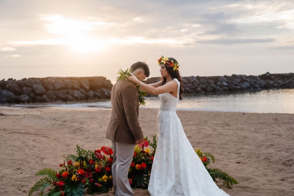 A bride and groom share a romantic moment on a beach during golden hour, with a floral arrangement visible in the foreground and calm water in the background.