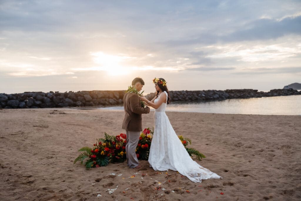 A couple shares an intimate moment on a sandy beach at sunset during their wedding ceremony, with floral decorations and calm waters in the background.