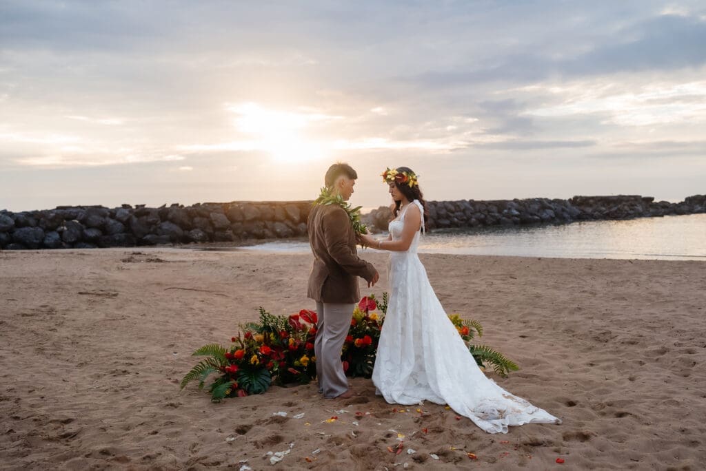 A bride and groom stand together on a sandy beach during golden hour, with a floral arrangement visible in the foreground and calm water in the background.