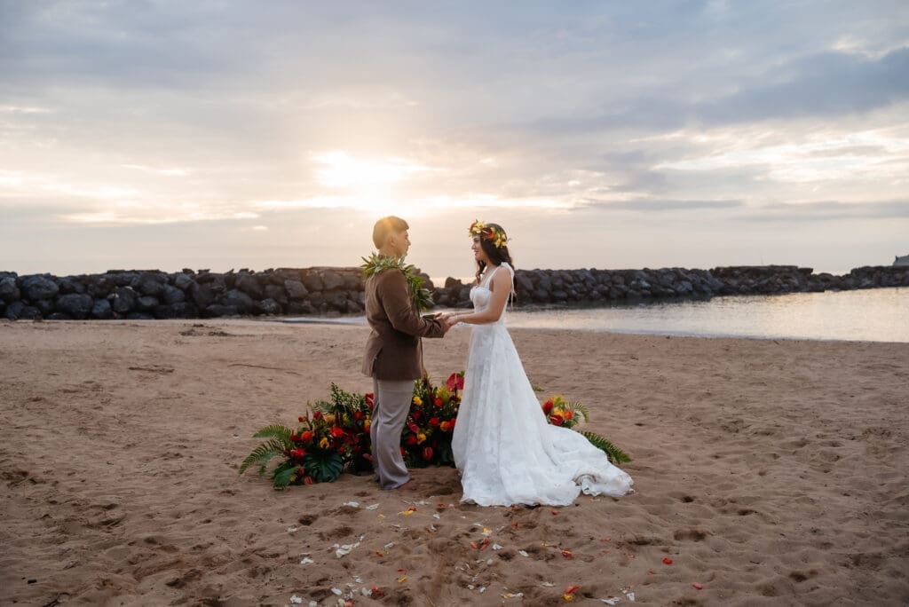 A bride and groom stand together on a sandy beach at golden hour, with floral decorations and a serene water backdrop creating a romantic wedding moment.