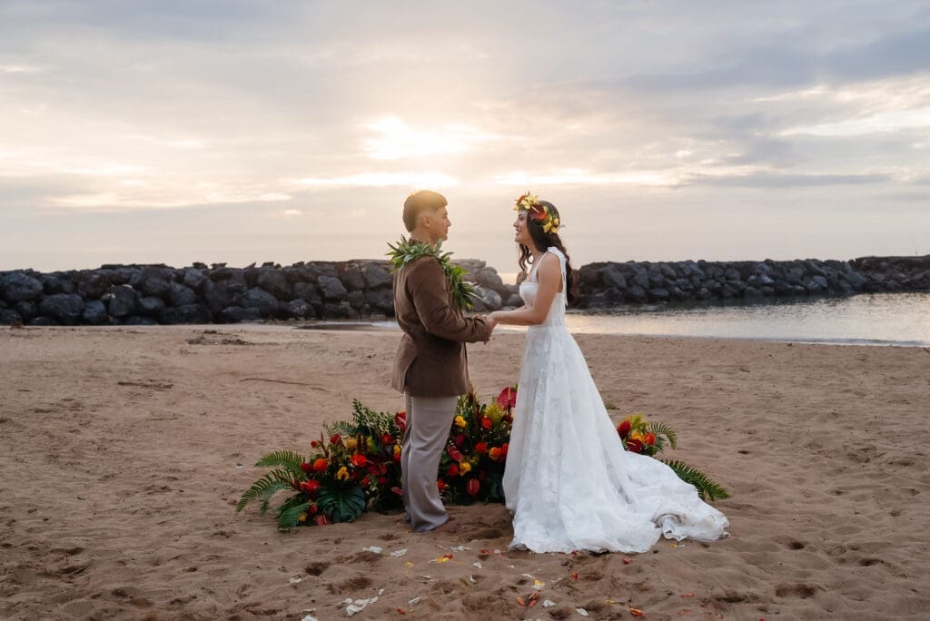 A couple in wedding attire shares a moment on a sandy beach at golden hour, with a floral arrangement visible nearby.