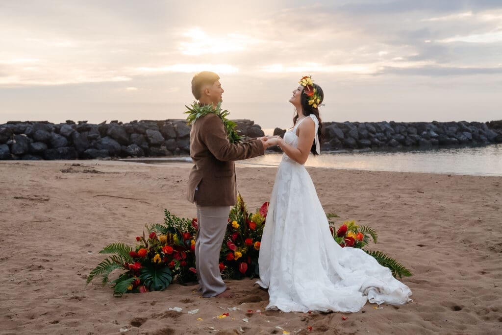 A bride and groom stand together on a sandy beach at golden hour, with a floral arrangement between them and water visible in the background.