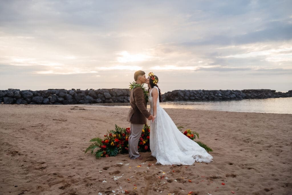 A bride and groom embrace on a sandy beach during golden hour with floral decorations and calm waters in the background.