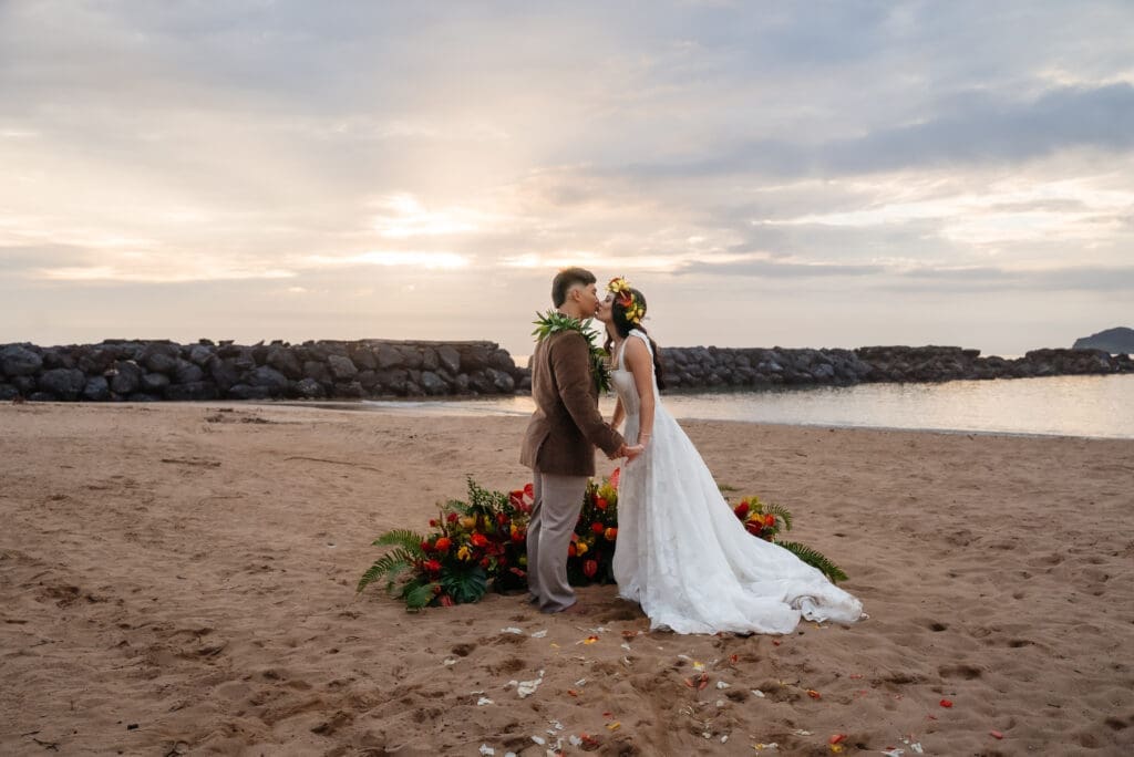A bride and groom pose together on a sandy beach during golden hour, with a floral arrangement and calm waters in the background.