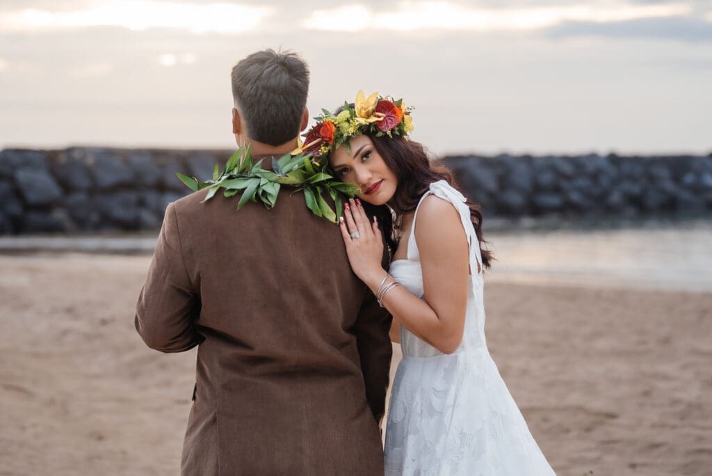 A bride and groom pose together on a beach during golden hour, with the groom facing away and the bride adorned with a lei garland necklace.