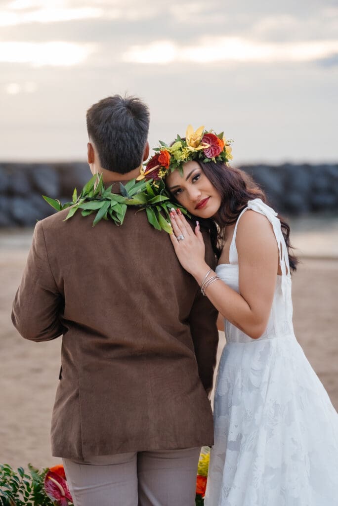 A bride wearing a white dress and colorful floral wreath embraces her groom in brown attire during golden hour near a waterfront.