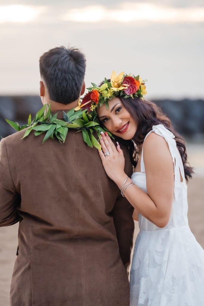 A couple in wedding attire embraces from behind, with the bride wearing a colorful flower lei around her neck, captured in an intimate close-up portrait.