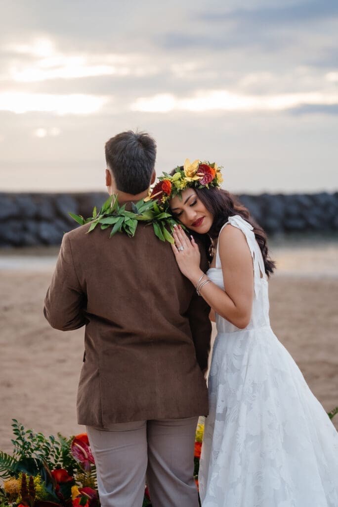 A bride and groom embrace on a beach during golden hour, with the bride wearing a flower crown and the groom in a brown suit, overlooking the water.