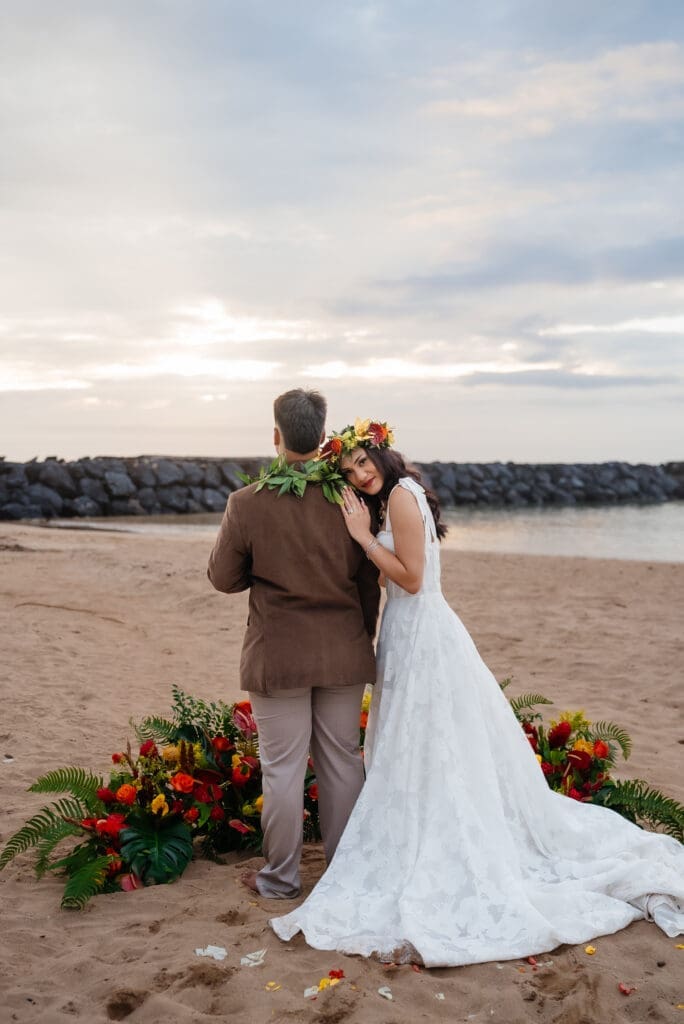 A bride and groom stand together on a sandy beach at golden hour, with floral arrangements beside them and a calm waterfront in the background.