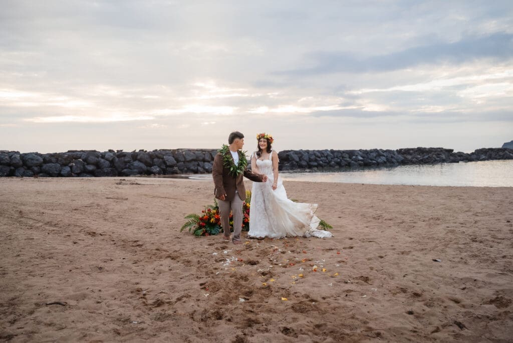A bride and groom stand together on a sandy beach at golden hour during their wedding ceremony, with calm waters and treeline visible in the background.