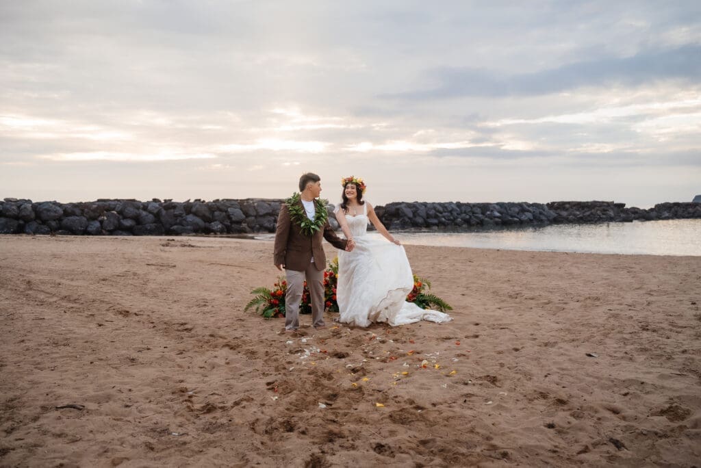 A bride and groom pose together on a sandy beach during golden hour with calm waters and cloudy skies in the background.