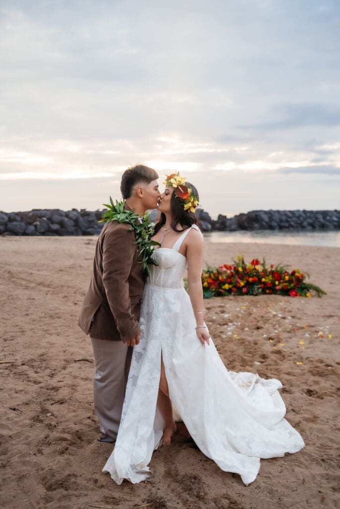 A bride and groom share an intimate moment on a sandy beach at golden hour, with the groom in a brown suit and the bride in a flowing white dress.