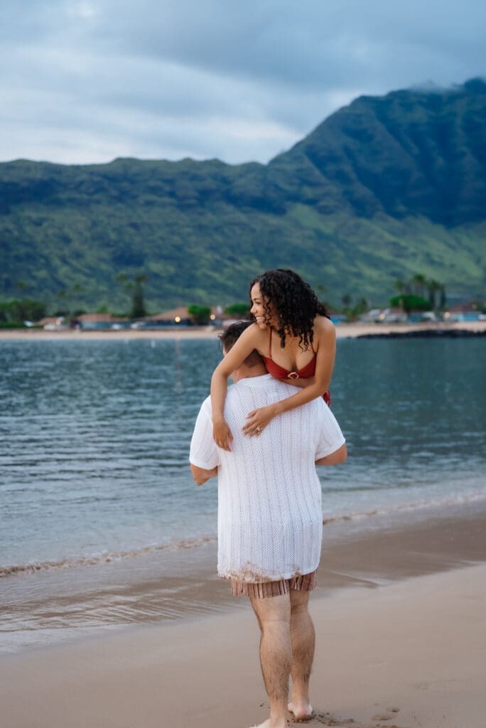 A pregnant woman in a white dress stands on a tropical beach with mountains in the background, cradling her belly in a serene maternity portrait.