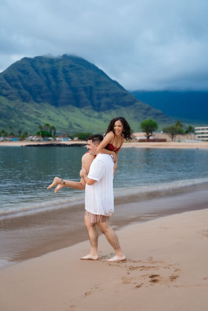 A woman in a white dress stands on a tropical beach with a dramatic mountain backdrop and calm blue ocean waters.